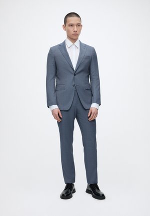 Mann steht, trägt einen slim-fit blauen Anzug, weißes Hemd und schwarze Anzugsschuhe auf einem schlichten weißen Hintergrund.