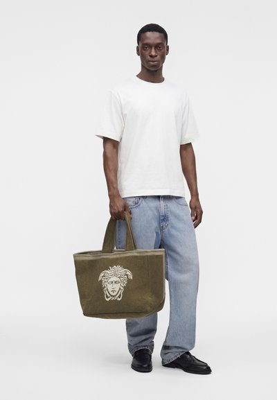 Homme portant un t-shirt blanc et un jean bleu clair tenant un sac fourre-tout vert olive avec un motif de tête de Méduse, debout sur un fond blanc uni.