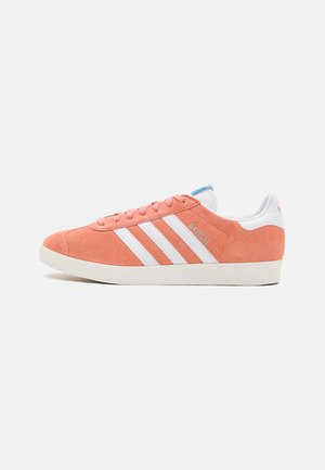Sneaker en suède orange avec des rayures et des accents blancs. Présente un col rembourré, des lacets plats et une semelle en caoutchouc texturé. Marque "Gazelle" notable.