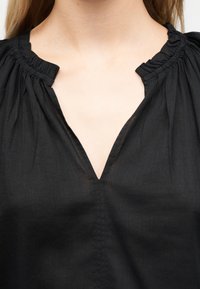 Κοντινό πλάνο του πάνω μέρους του στήθους ενός ατόμου που φοράει μαύρο μπλουζάκι με βολάν στο λαιμό και μπροστινό άνοιγμα σε σχήμα V.