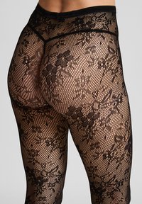 Zwarte kanten leggings met een bloemmotief, voorzien van een mesh-ontwerp en een elastische tailleband, weergegeven vanaf de achterkant, met een nadruk op textuur en detail.