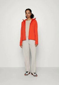 Woman standing against plain background wearing beige pants, beige top, bright orange zip-up jacket, and white sneakers with orange and black details.