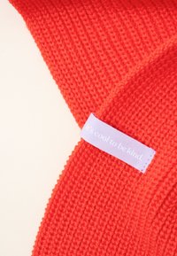 Red knitted accessory with a ribbed texture. Features a light purple label with text “It's cool to be kind.”