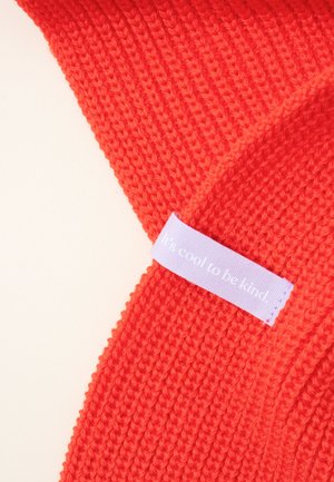 Red knitted accessory with a ribbed texture. Features a light purple label with text “It's cool to be kind.”