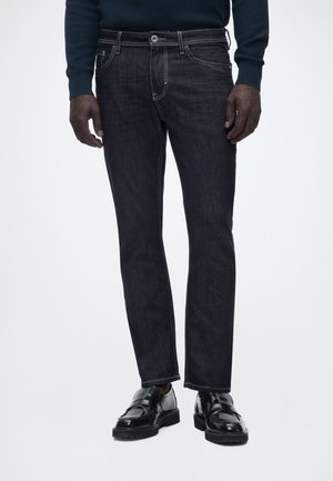 Mand iført mørkeblå jeans, sorte læderloafers og en mørk langærmet top, stående mod en ensfarvet baggrund.