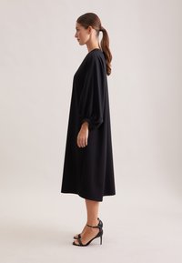 Schwarzes, knielanges Kleid mit lockerer Passform, puffärmeligen Ärmeln und weicher Textur. Getragen mit schwarzen, hochhackigen Sandalen.