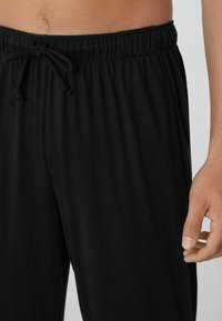 Schwarze Jogginghose mit einem elastischen Bund, glatter Textur und lässiger Passform, bei der ein Teil einer Hand am Bund sichtbar ist.