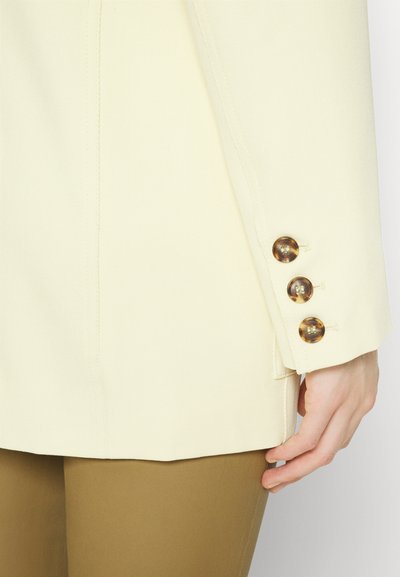 Κοντινό πλάνο ενός μπεζ σακακιού με τρία κουμπιά από φολιδωτό υλικό, που φοριέται πάνω από καφέ παντελόνι, με το χέρι να ακουμπά κοντά στο ισχίο.