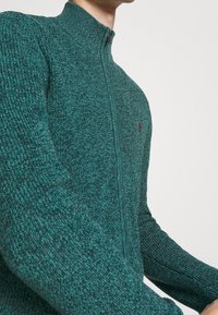 Zip-up tröja i strukturerad tealstickning, med hög krage och en liten röd logotyp. Tätsittande ärmar med ribbade muddar.