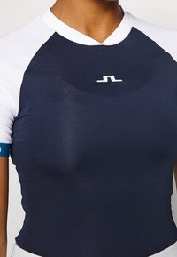 Marinblå atletisk topp i en figurnära design, med vita ärmar och en liten logotyp på bröstet. Slät, stretchig tyg.