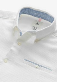 Polo shirt blanc avec une surface texturée, col intérieur à motif bleu clair, et une petite poche sur la poitrine avec des détails assortis. Les boutons sont blancs.