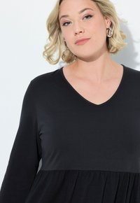 Schwarzes, langärmeliges Kleid mit V-Ausschnitt und gerüschtem Rock. Der Stoff ist glatt und weich, was eine definierte Silhouette schafft.