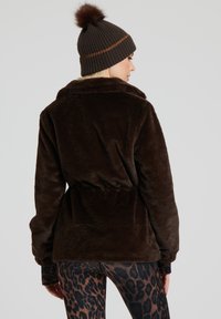 Brun fuskpälsjacka med bred krage och bälte i midjan, bärs över mönstrade leggings, kompletterad med en ribbad brun mössa med pom-pom.