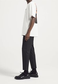 Homme portant un t-shirt blanc à motif, un pantalon noir raccourci, des chaussettes noires et des chaussures habillées noires à lacets, debout de profil.