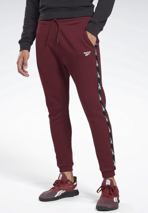 Hombre con pantalones deportivos Reebok burdeos con una franja de logo en negro y blanco a los lados, combinados con zapatillas deportivas burdeos y negras a juego.