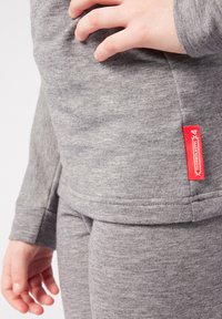 Felpa grigia e pantaloni realizzati in un tessuto morbido; presenta un'etichetta rossa con testo. Ingrandimento di una mano appoggiata sulla vita.