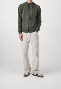 Felpa verde con cappuccio e zip, dotata di tasche frontali e logo. Abbinata a pantaloni beige chiaro e sneakers grigie con accenti neri.