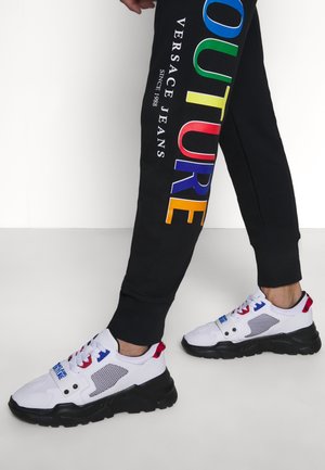 Persona che indossa pantaloni neri con scritta colorata "COUTURE VERSACE JEANS" e sneakers bianche chunky con suole nere e dettagli rossi e blu.