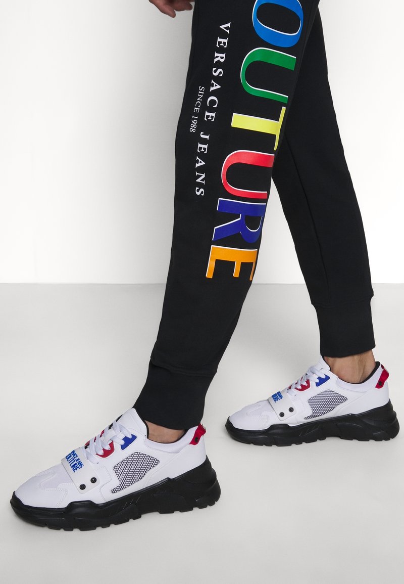Personne portant un pantalon noir avec le texte coloré "COUTURE VERSACE JEANS" et des baskets blanches épaisses avec des semelles noires et des touches rouges et bleues.