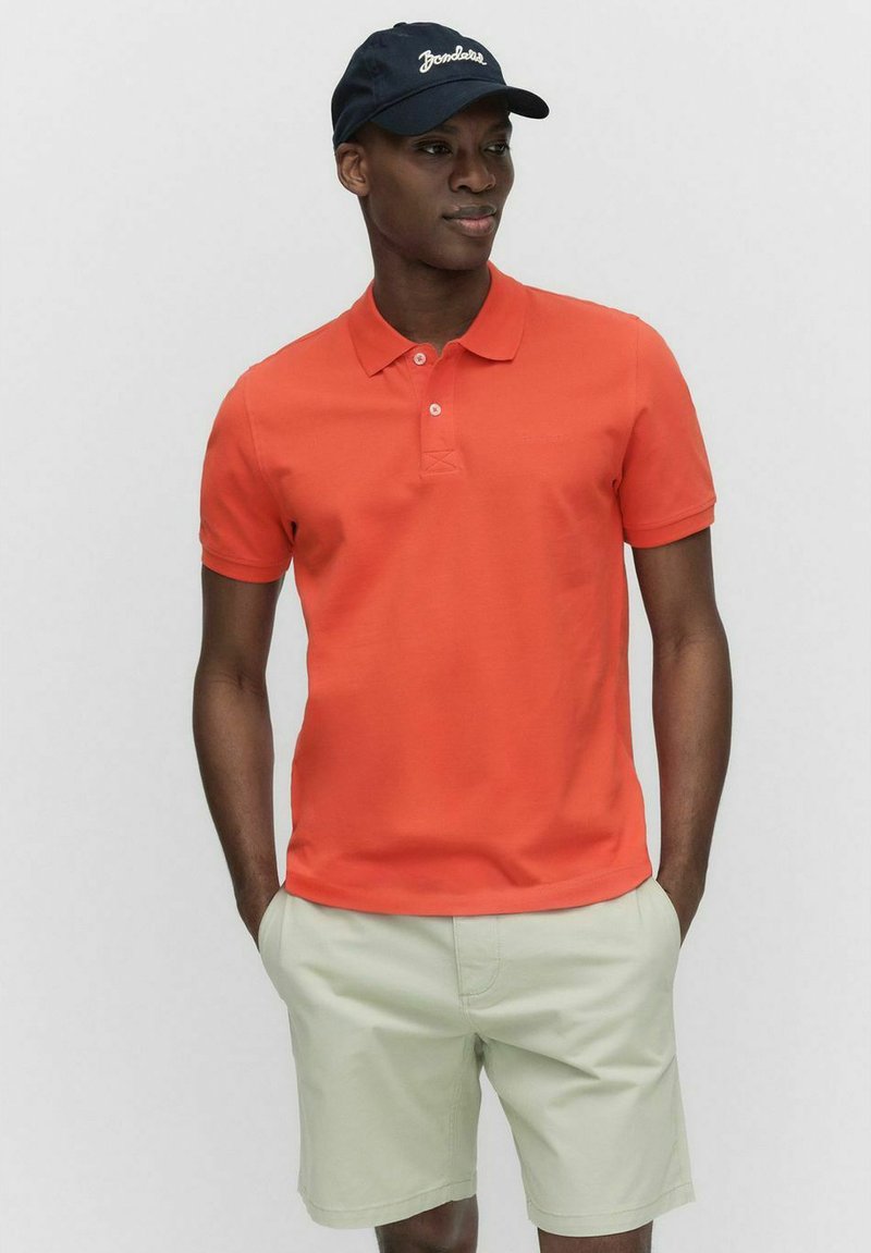 Orange polo shirt med krave; korte ærmer. Bæres med lysegrønne shorts. Modellen bærer også en marineblå kasket med hvid tekst.