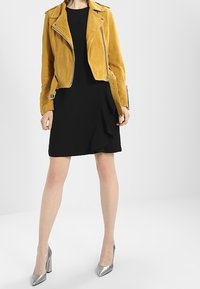 Giacca da moto in suede gialla indossata sopra un vestito nero con orlo a volant; abbinata a scarpe con tacchi alti argento.