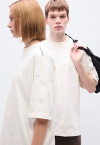 Κρεμ πουκάμισα με κοντά μανίκια, με ανάγλυφα σχέδια και λογότυπο στον ώμο. Ένα μοντέλο κρατάει ένα μαύρο σακάκι. Ομαλή υφή.