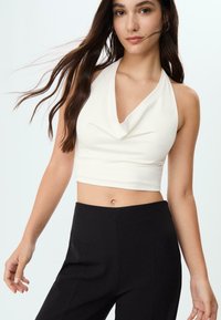 Bela crop majica s halter vratom in detajlom od tkanine, v kombinaciji s črnimi hlačami visoko pasu, ki poudarjajo gladko tkanino in oprijet silhueto.