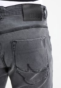 Šedé džíny z denimu s opotřebovanou strukturou, s černou koženou nášivkou na pase a standardním designem zadní kapsy.
