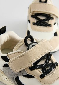 Baskets pour enfants beige et noires avec une tige texturée, une bande Velcro et des lacets noirs. Comprend des accents gris et une semelle rembourrée.