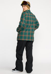 Chemise en flanelle à carreaux verts avec des lignes brunes et orange, pantalon cargo noir et bottes de snowboard noires, accompagnés d'un béret marron et de lunettes de protection.
