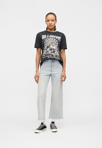 Νεαρό άτομο με πλεξούδες στα μαλλιά, φορώντας μαύρο t-shirt με σχέδιο, ανοιχτό μπλε καλλιτεχνικό τζιν, λευκές κάλτσες και μαύρα αθλητικά παπούτσια, σε απλή φόντο.