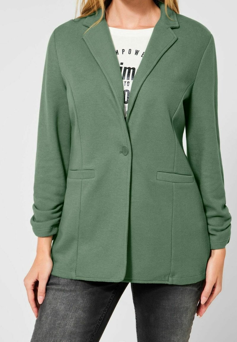 Zelené sako z měkké tkaniny, s výřezovým límcem, zapínáním na jeden knoflík a bočními kapsami. Střih s ohrnutými rukávy.
