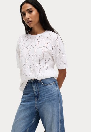 Hvid bomulds-t-shirt med cirkulære udskæringsmønstre, korte ærmer og afslappet pasform, parret med højhalsede blå denimjeans.