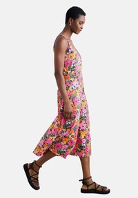 Robe d'été à motifs floraux présentant un mélange vibrant de rose, orange et vert. Tissu doux, longueur cheville avec de larges bretelles, accompagnée de sandales noires.