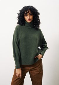 Groene gebreide coltrui met een ontspannen pasvorm, geribbelde boorden en onderrand, gecombineerd met bruine broek. Zachte textuur en casual ontwerp.