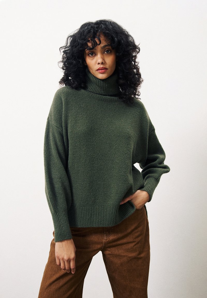 Groene gebreide coltrui met een ontspannen pasvorm, geribbelde boorden en onderrand, gecombineerd met bruine broek. Zachte textuur en casual ontwerp.