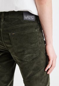 Zaļas kordveida bikses ar vertikālajām ribām, ar melnu ādas etiķeti jostas vietā un aizmugurējo kabatu ar mazu zīmola birku.