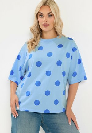 Frau mit blondem Haar, die ein hellblaues oversized T-Shirt mit dunkelblauen Punkten und blaue Jeans trägt, posiert vor weißem Hintergrund.