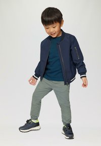 Marine bomberjack met een ritssluiting aan de voorkant en een zijzak, gedragen met een teal gestreept shirt en lichtgroene broek, en zwarte sneakers.