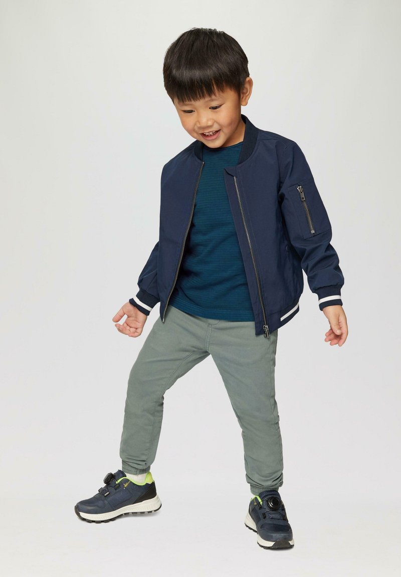 Marine bomberjack met een ritssluiting aan de voorkant en een zijzak, gedragen met een teal gestreept shirt en lichtgroene broek, en zwarte sneakers.