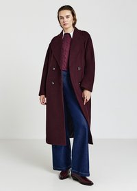 Cappotto doppiopetto color bordeaux con ampi revers e bottoni metallici, abbinato a un maglione girocollo color prugna con trecce e pantaloni di jeans a vita alta e gamba svasata.