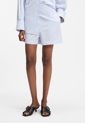 Personne portant un short rayé bleu clair et blanc et une chemise assortie à manches longues, avec des sandales noires à talons bas avec des boucles.