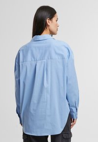 Camicia button-up oversize azzurro chiaro con piega sul retro, colletto classico e maniche lunghe. Realizzata in un tessuto liscio e resistente alle pieghe.