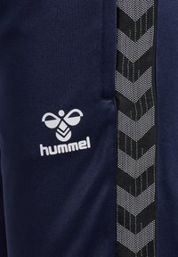Marineblauwe broek met een gladde textuur, voorzien van een zwarte zijpaneel met een chevronpatroon en een wit "hummel" logo.