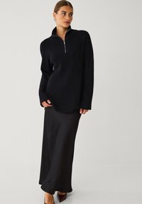 Sort ribbet strikket pullover med lynlås-krave, parret med en elegant sort maxi-nederdel. Ærmerne er brede, og stoffet har en struktureret overflade.