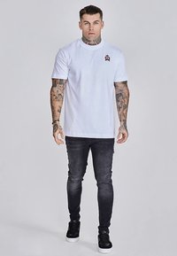 Weißes Baumwoll-T-Shirt mit Rundhalsausschnitt und kurzen Ärmeln, versehen mit einem kleinen gestickten Logo. Kombiniert mit dunkelgrauen Slim-Fit-Jeans und schwarzen Turnschuhen.