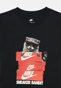 Ρακούν με γυαλιά ηλίου κοιτάζει από πάνω τριών στοιβαγμένων κόκκινων κουτιών παπουτσιών Nike, με το κείμενο "Sneaker Bandit" από κάτω σε μαύρη μπλούζα.