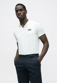 Polo azul claro com gola e mangas curtas, apresentando um logótipo preto "KARL LAGERFELD" no peito esquerdo. Usado com calças azuis escuras.