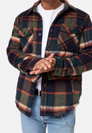Mand iført en knaplukket mørkegrøn, orange og beige ternet jakke over en hvid skjorte og lyseblå jeans, hænderne foldet foran.