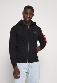 Black zip-up hoodie with a kangaroo pocket, hood, and a logo patch on the chest. Paired with light blue jeans.